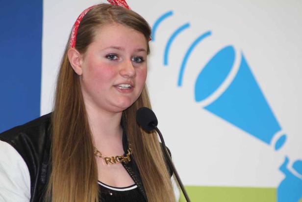 Eine junge Frau mit rotem Haarband spricht in ein Mikrofon vor einem blauen Lautsprecher-Logo.