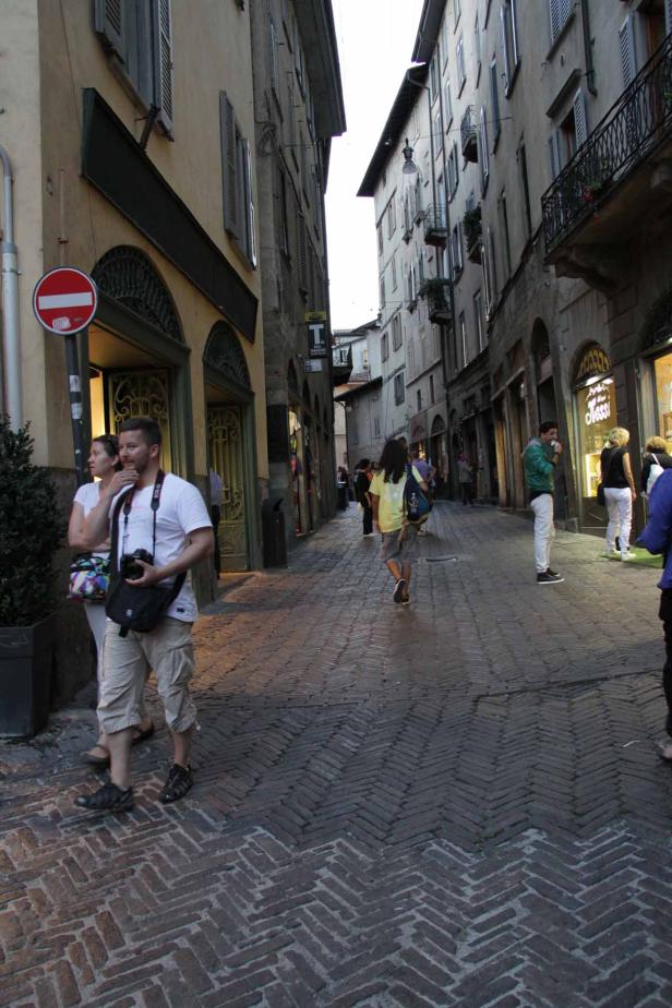Menschen gehen eine schmale Gasse mit Kopfsteinpflaster entlang.