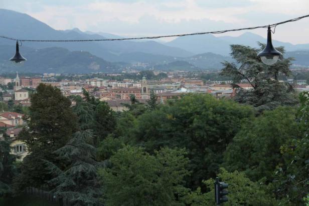 Blick über eine Stadt mit Bergen im Hintergrund, gerahmt von Bäumen und zwei Laternen.