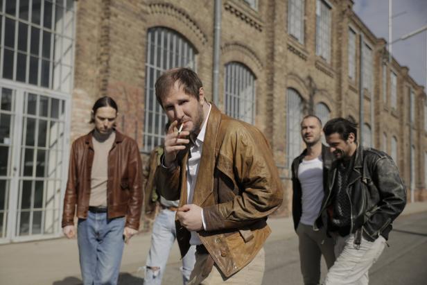 Eine Gruppe von Männern in Lederjacken steht auf der Straße, einer raucht eine Zigarette.
