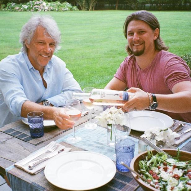 Zwei Männer sitzen an einem Tisch im Freien, während einer Wein einschenkt.