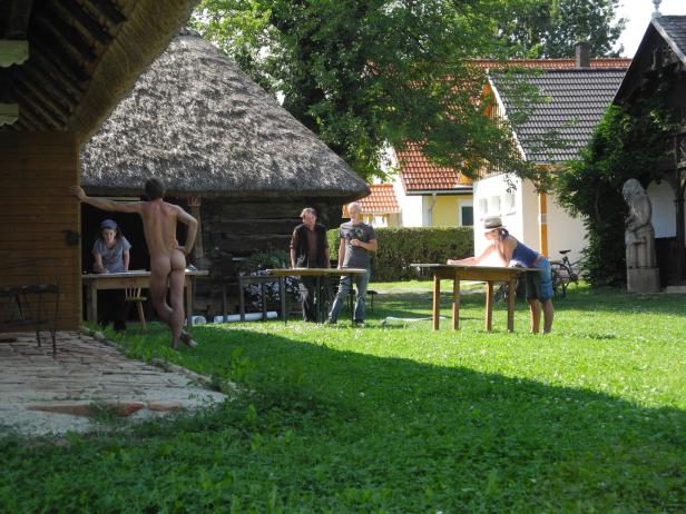 Eine Gruppe von Menschen steht im Freien an Tischen, eine nackte Person im Vordergrund.