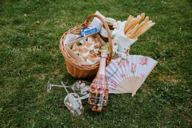 Ein Picknickkorb mit Wein, Gläsern, Gebäck und einem Fächer auf einer Wiese.