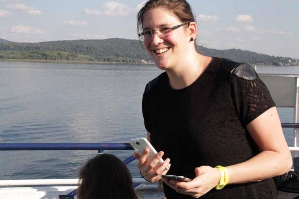 Eine lächelnde Frau mit Brille hält zwei Smartphones vor einem See.