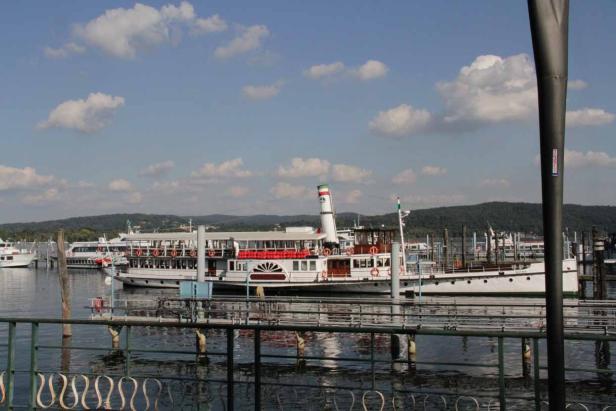Ein Schaufelraddampfer liegt an einem See vor bewaldeten Hügeln.