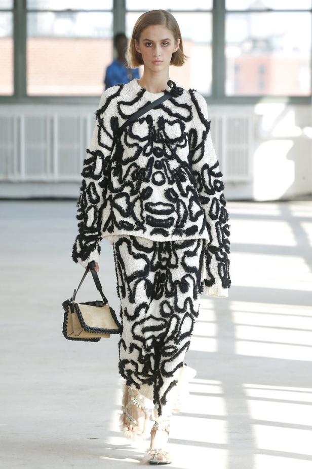 Ein Model präsentiert ein weißes Ensemble mit schwarzen, abstrakten Mustern und eine Handtasche.