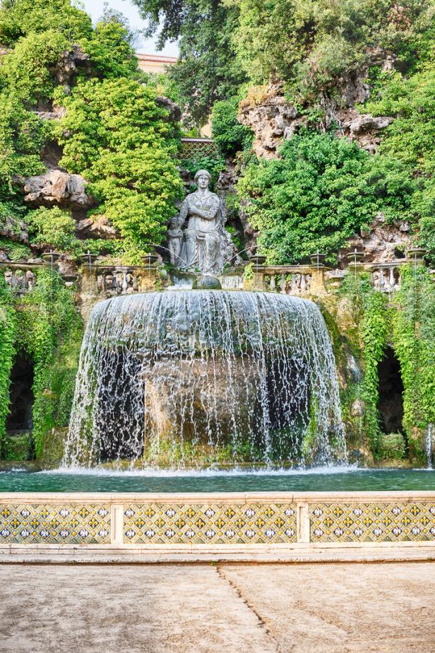 Ein Wasserfall mit einer Statue im Hintergrund, umgeben von üppigem Grün.