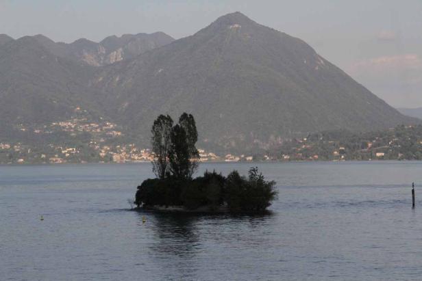 Eine kleine, bewaldete Insel liegt in einem See vor einer Bergkulisse.