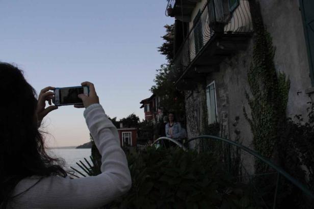 Eine Person fotografiert mit einem Smartphone eine Frau vor einem Haus am See.