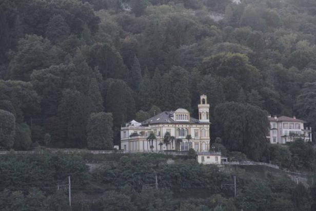 Ein großes, gelbliches Gebäude mit Turm steht auf einem bewaldeten Hügel.