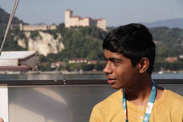 Ein junger Mann mit gelbem T-Shirt und Ausweis vor einer Landschaft mit See und Burg.