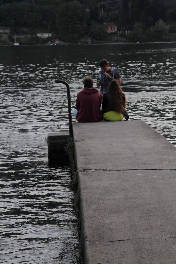 Drei Personen sitzen auf einem Steg am Wasser.
