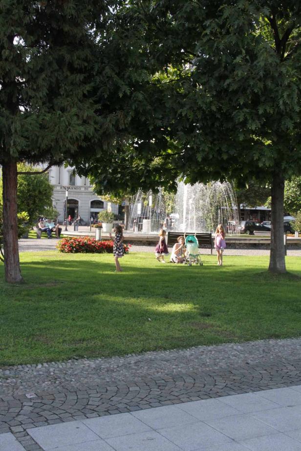 Eine Frau mit Kindern entspannt sich auf einer Wiese vor einem Springbrunnen.