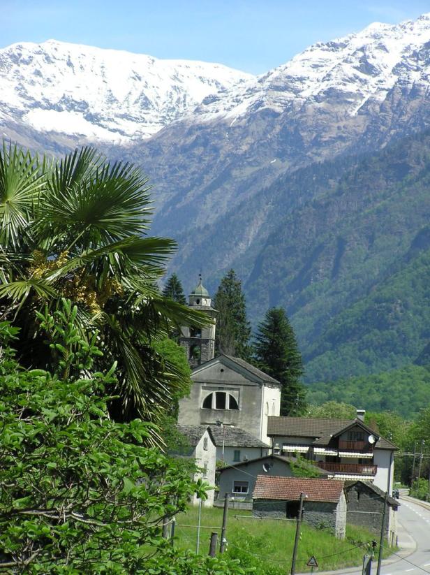 Eine Kirche und Häuser liegen vor einer bewaldeten Berglandschaft mit schneebedeckten Gipfeln.