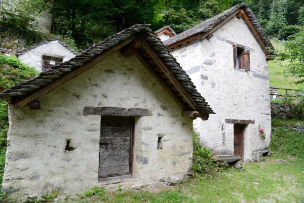 Drei kleine, steinerne Häuser mit dunklen Holztüren und Schieferdächern stehen im Grünen.