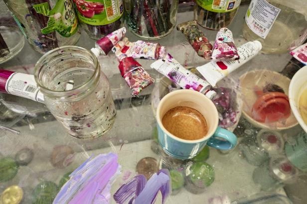 Ein chaotischer Tisch mit Farbtuben, Gläsern, Pinseln und einer Tasse Kaffee.