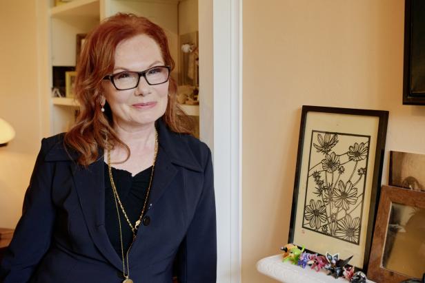 Eine Frau mit roten Haaren, Brille und dunklem Blazer steht neben einem Regal mit Dekorationen.