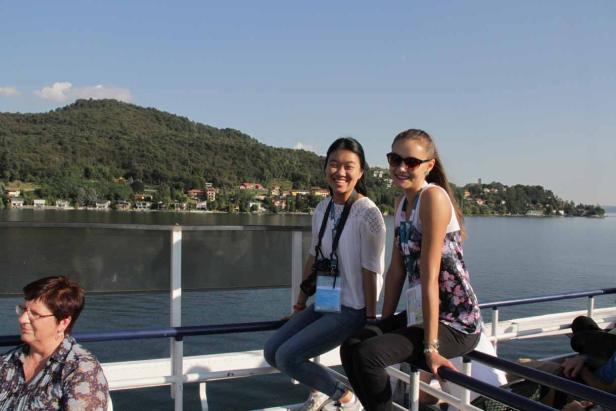 Zwei junge Frauen sitzen auf einer Fähre vor einer bewaldeten Hügellandschaft.