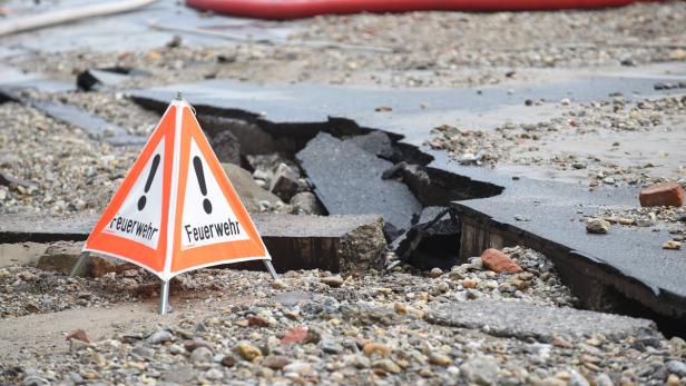 Ein orangefarbenes Warnschild der Feuerwehr steht vor einer zerstörten Straße.