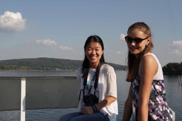 Zwei junge Frauen sitzen an einem See und lächeln.