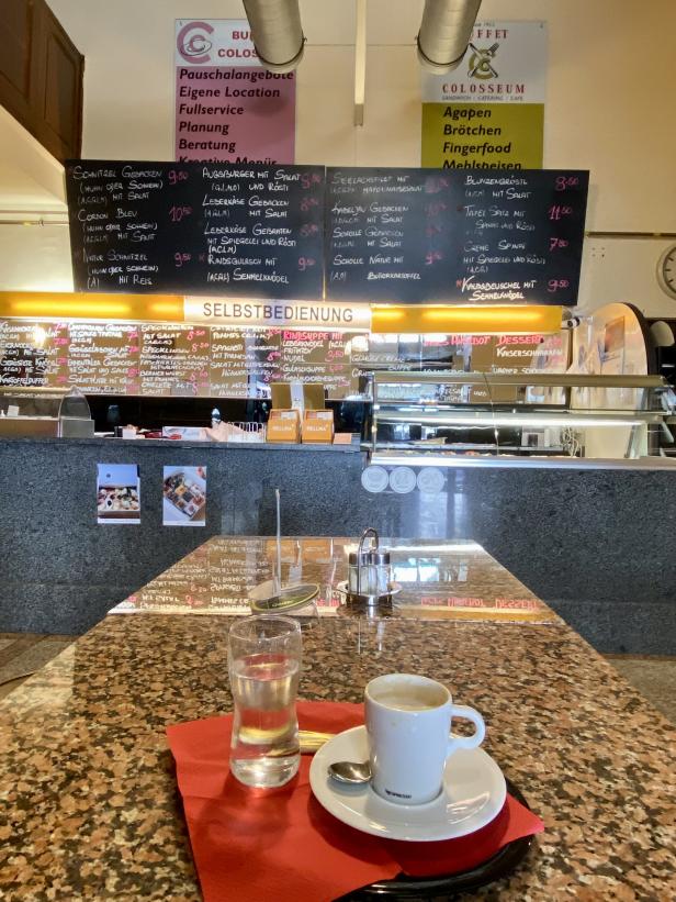 Ein Espresso und ein Glas Wasser stehen auf einem Tisch in einem Café mit Speisekarte im Hintergrund.