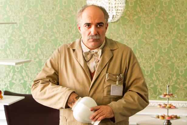 Ein Mann mit Schnurrbart hält eine weiße Schale in einem Raum mit gemusterter Tapete.