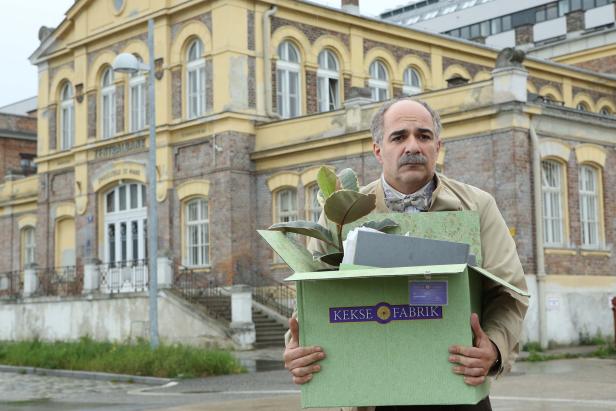 Ein Mann mit Fliege trägt einen Karton mit der Aufschrift „Kekse Fabrik“ vor einem Gebäude.