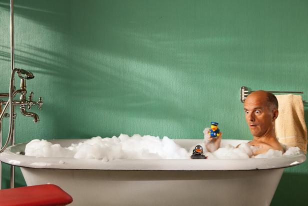 Ein Mann sitzt überrascht in einer mit Schaum gefüllten Badewanne und spielt mit Quietscheentchen.
