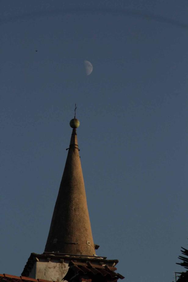 Ein Kirchturmspitz ragt vor einem blauen Himmel mit einer Mondsichel auf.