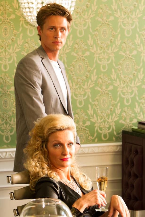 Eine Frau sitzt mit einem Glas Sekt, während ein Mann im Hintergrund steht.