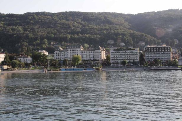 Blick über den See auf eine Stadt am Ufer mit bewaldeten Hügeln im Hintergrund.