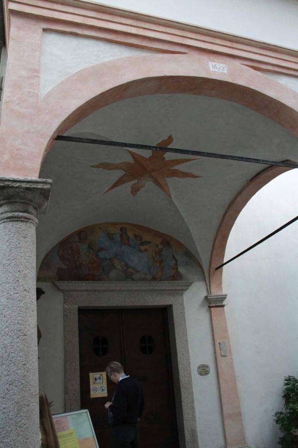 Ein Mann betrachtet eine Informationstafel vor einem Gebäude mit Malereien und einer Jahreszahl über dem Eingang.