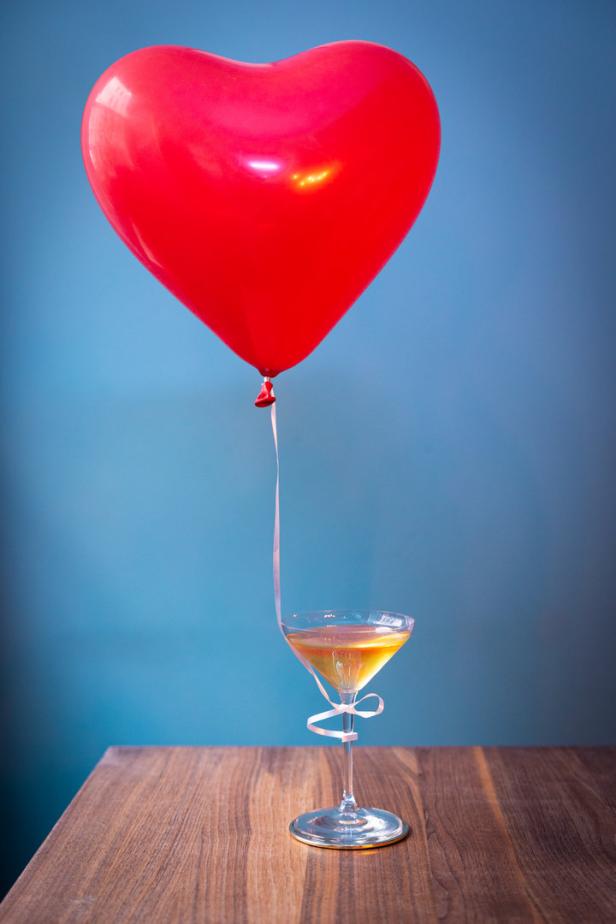 Ein roter Herzluftballon schwebt über einem Cocktailglas auf einem Holztisch.