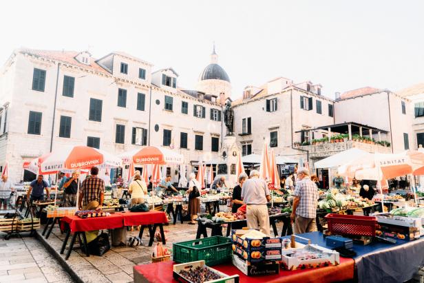 Ein Marktplatz in Dubrovnik mit Verkaufsständen unter Sonnenschirmen vor historischen Gebäuden.