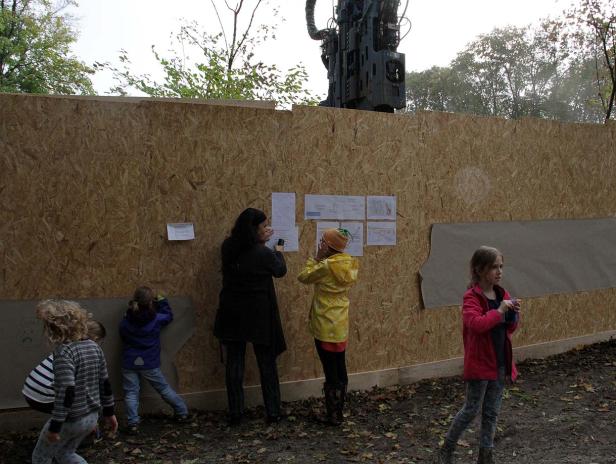 Eine Gruppe von Kindern und eine Frau betrachten Pläne, die an einer Holzwand befestigt sind.