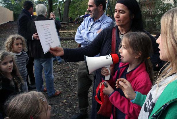 Eine Frau hält ein Megafon, während ein Mädchen spricht; andere Kinder und Erwachsene stehen um sie herum.