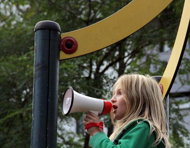 Kids, hockegasse Ein blondes Mädchen spricht in ein Megaphon auf einem Spielplatz.