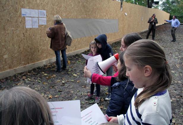 Eine Gruppe Kinder protestiert mit einem Megafon und Plakaten vor einer Holzwand.