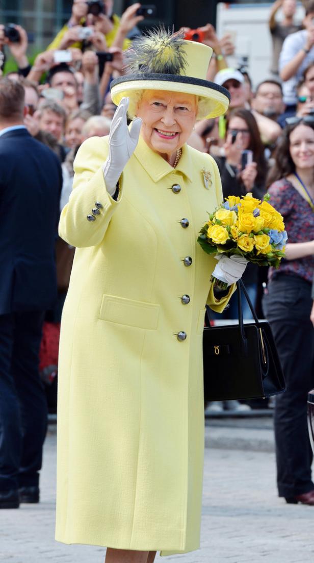 Königin Elisabeth II. winkt mit einem gelben Blumenstrauß in der Hand einer Menschenmenge zu.