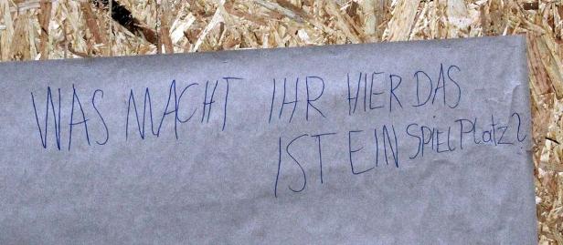 Ein Zettel an einer Holzwand fragt: „Was macht ihr hier das ist ein Spielplatz?“.
