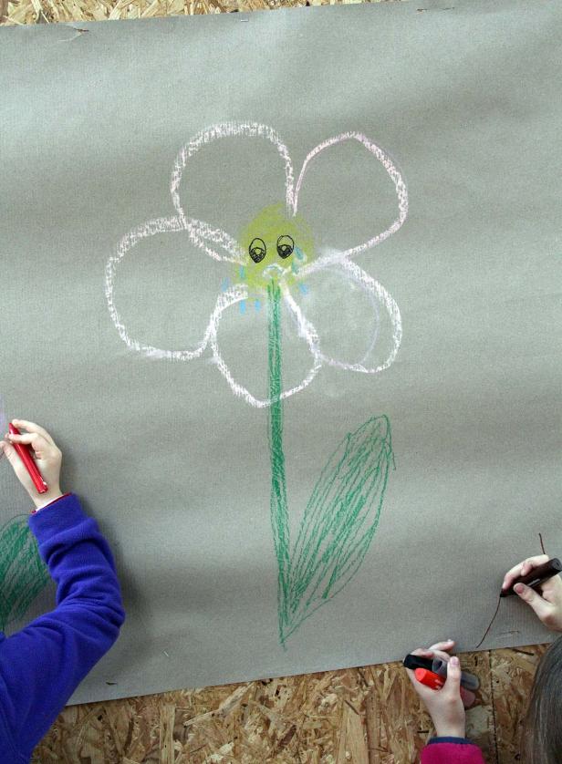 Kinder zeichnen mit Wachsmalstiften eine Blume mit traurigem Gesicht auf Papier.