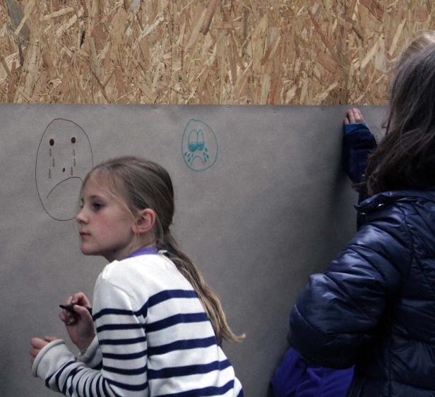 Zwei Mädchen zeichnen traurige Gesichter auf eine graue Leinwand.
