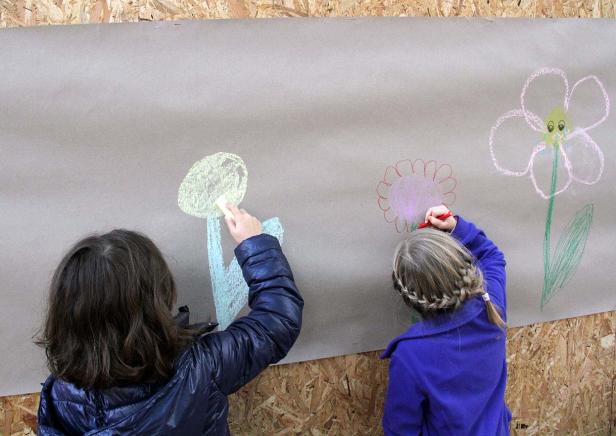 Zwei Kinder malen mit Wachsmalstiften Blumen auf ein großes Blatt Papier.