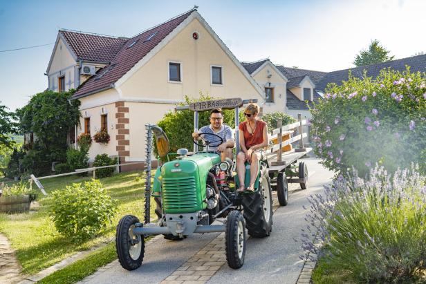 Ein Mann und eine Frau fahren mit einem grünen Traktor mit Anhänger durch eine ländliche Gegend.