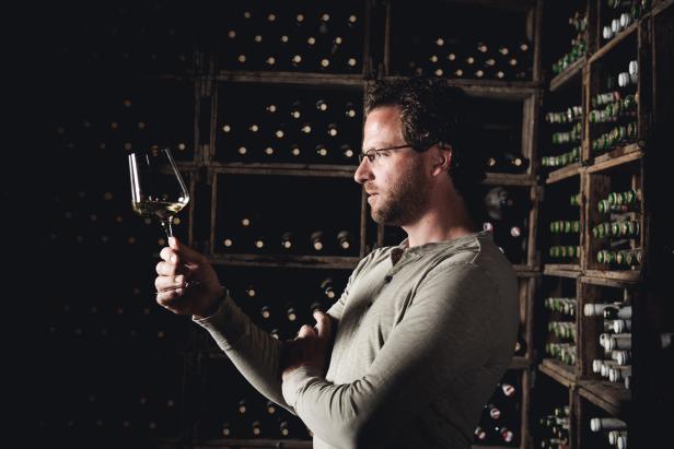 Ein Mann mit Brille betrachtet ein Glas Weißwein vor einem Regal voller Weinflaschen.