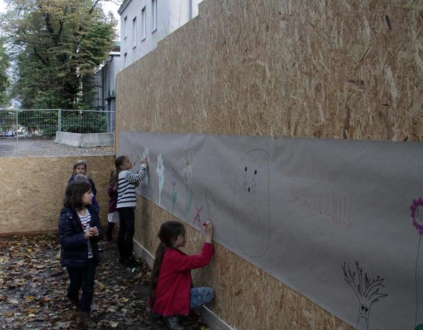 Mehrere Kinder malen auf einem langen Papier, das an einer Holzwand befestigt ist.