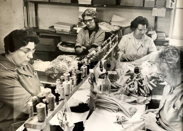 In einer Fabrik arbeiten vier Frauen an einer Werkbank mit Nähgarn und Stoffen.