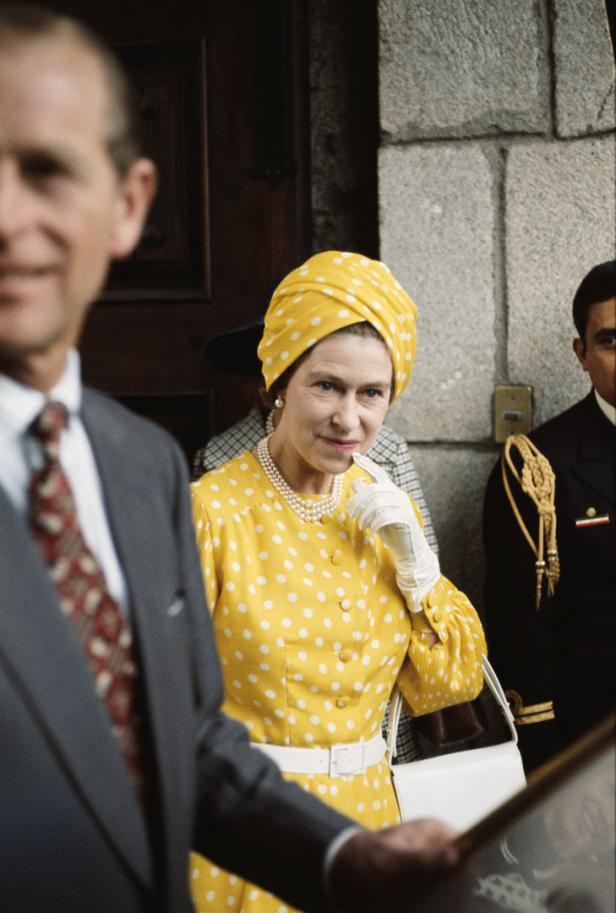 Königin Elisabeth II. trägt ein gelbes Kleid mit weißen Punkten und einen Turban.