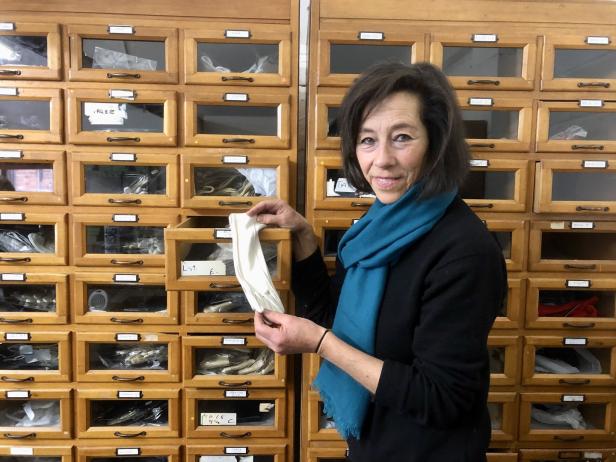 Eine Frau hält einen weißen Stoffhandschuh vor einem Schrank mit vielen Schubladen.