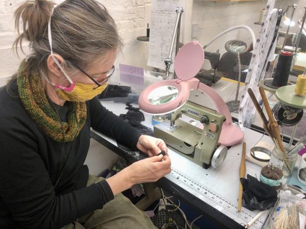 Eine Frau mit Maske arbeitet an einem Tisch mit einer Nähmaschine und einer Lupe.
